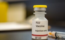 A vial of flu vaccine with a syringe at the background
